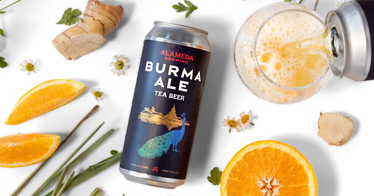 A can of Alameda Brewing Burma Ale surrounded by chamomile flowers, lemongrass stalks, fresh ginger, and orange slices, with a glass being poured alongside.