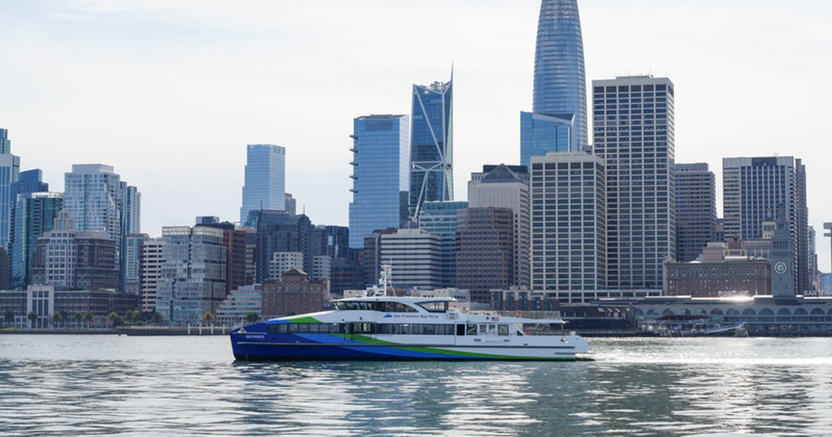 An SF Bay Ferry vessel in its blue and green livery crossing San Francisco Bay with the city skyline behind it.